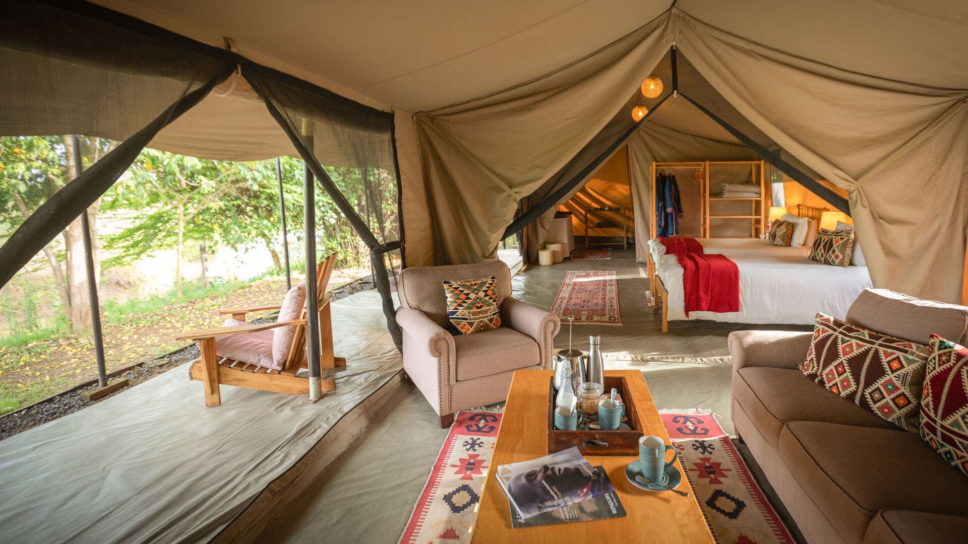 Interior of a large safari tent with seating area, beds, and open sides overlooking greenery.